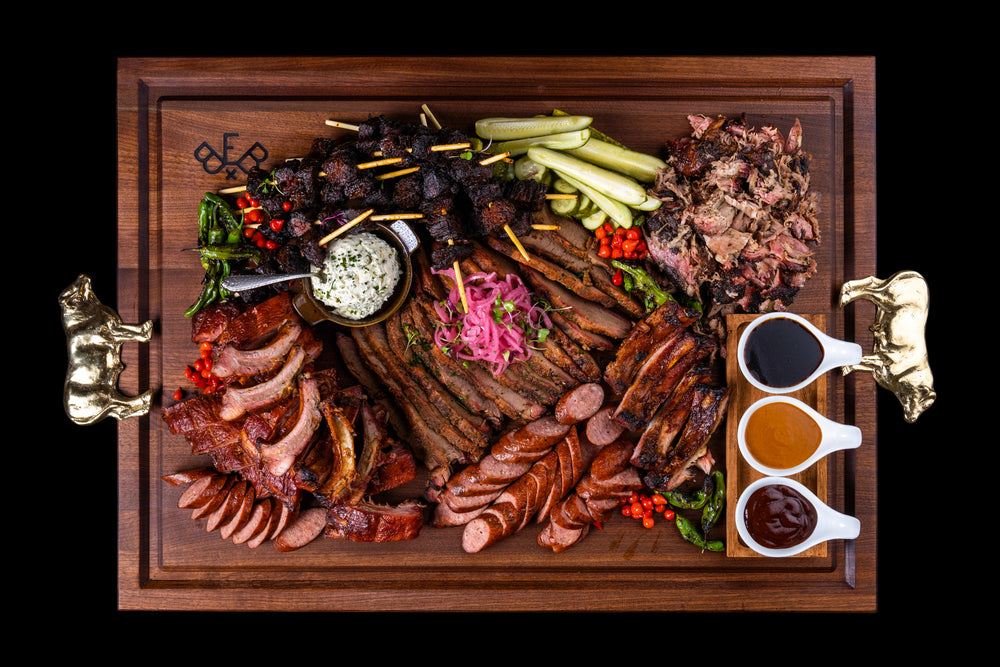 Pitmaster board in walnut loaded with a full spread of burnt ends, sliced brisket, sausage, pulled pork, and three sauces